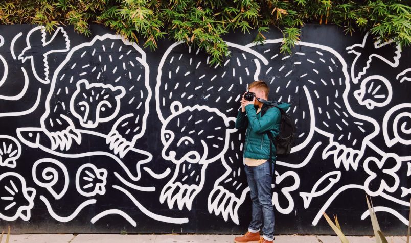 Young man holding a camera standing in front of a mural