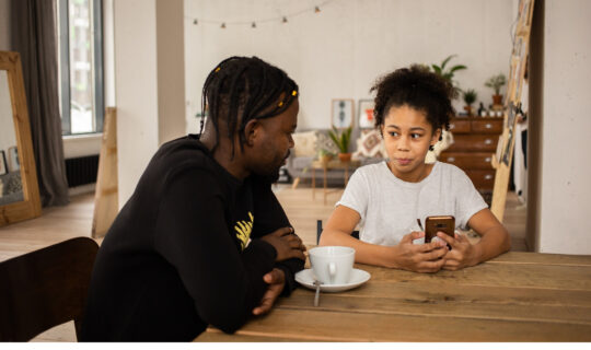 Two people at table, kitchen, holding phone, parent and child, discussing